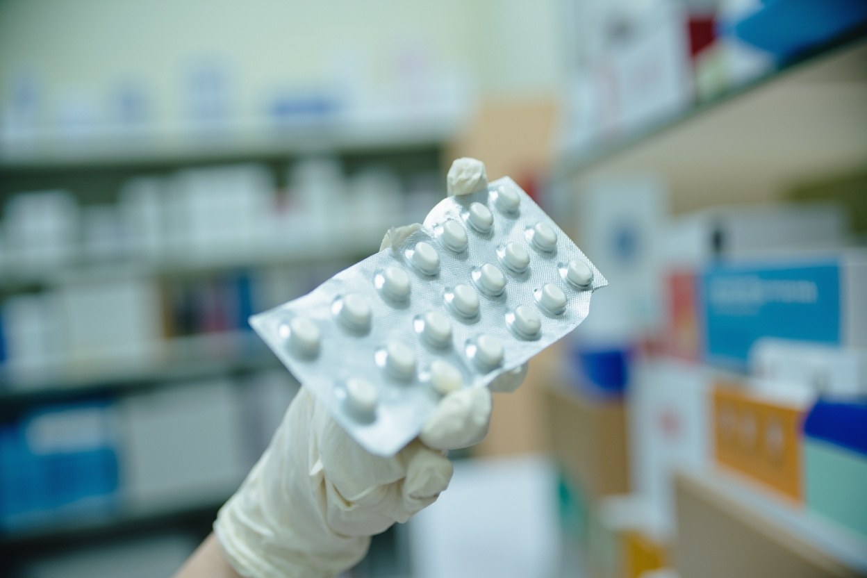 A close-up of a pharmacist's gloved hand holding a silver blister pack against a pale dispensary interior.