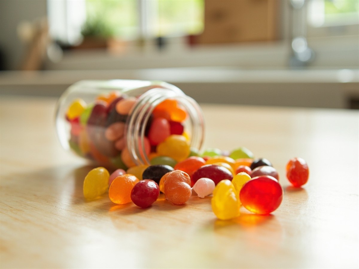 A small glass jar of glossy jelly beans tipped onto a kitchen counter, one cut in half showing the chewy centre.